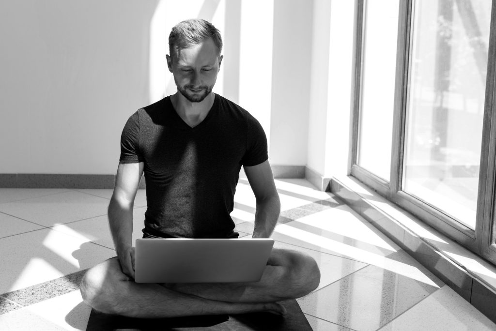 Young bearded man sitting on his yoga mat checking his laptop
