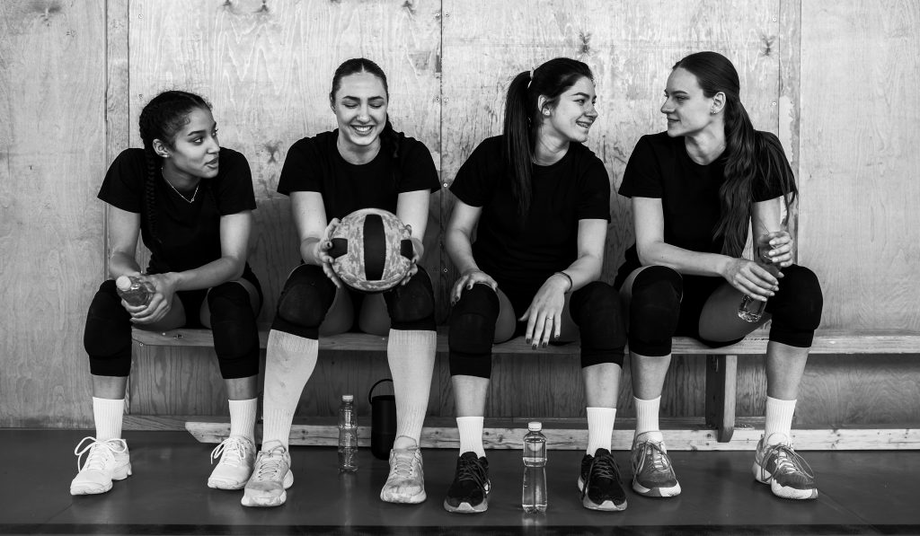 Portrait of a teenage female volleyball team is sitting on a break on bench on volleyball court and smiling. A multicultural young sportswomen are sitting on a bench and taking a break after training.