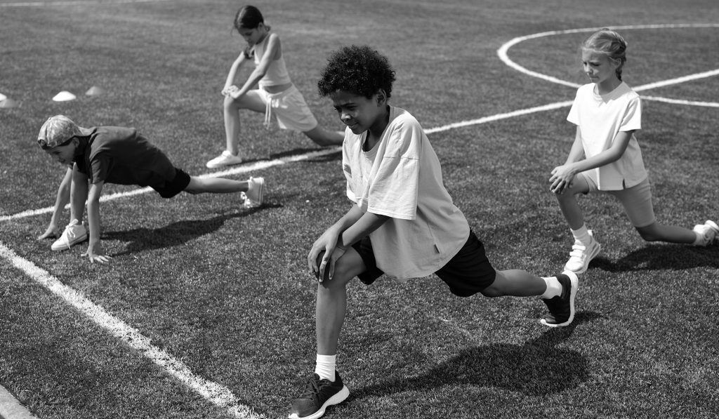 School boy in activewear doing stretching exercise on grass at stadium while his classmates repeating after him